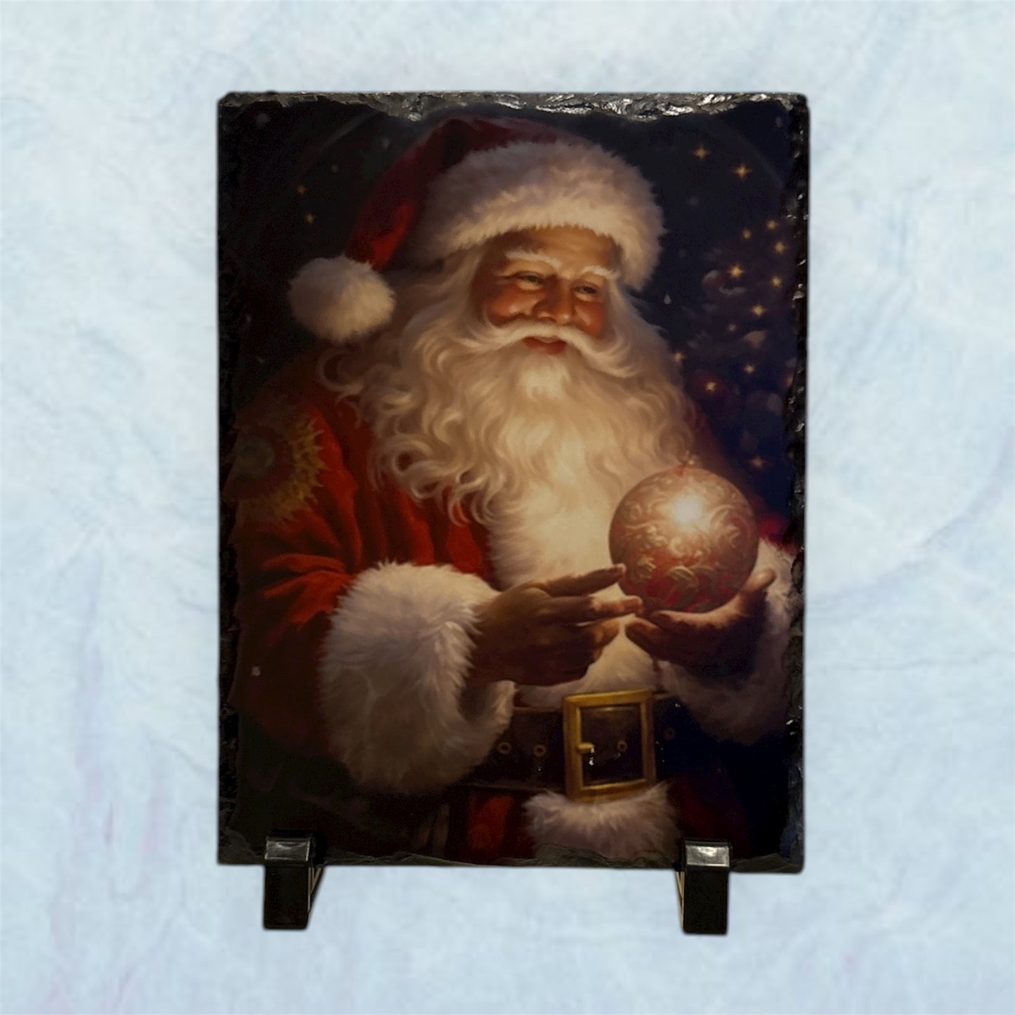 A rectangular slate plaque featuring a painted portrait of Santa Claus in his red suit holding a glowing ornament, displayed on a small black stand against a light blue textured background.